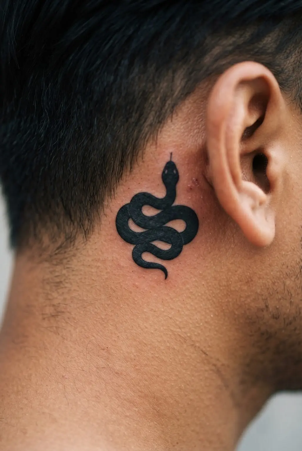 Coiled snake head tattoo behind the ear, showing a dark silhouette, curved body, pointed snout, and small eye detail.