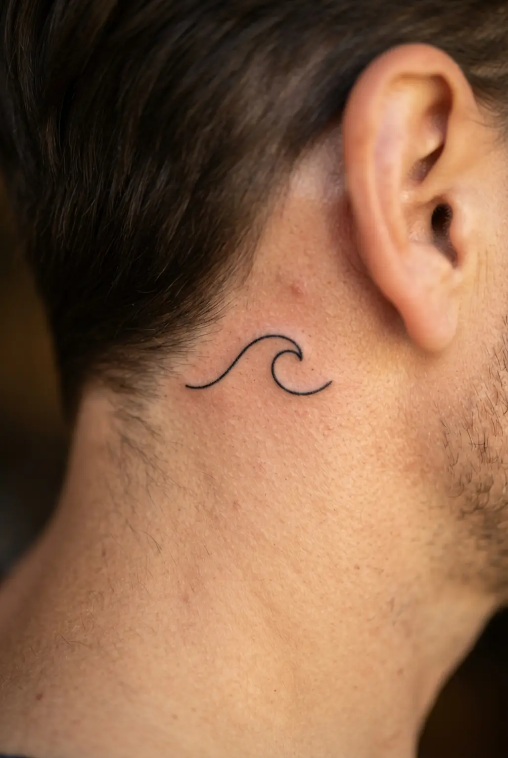 Single-line black wave crest tattoo behind the ear, with one continuous curve forming a compact ocean symbol.