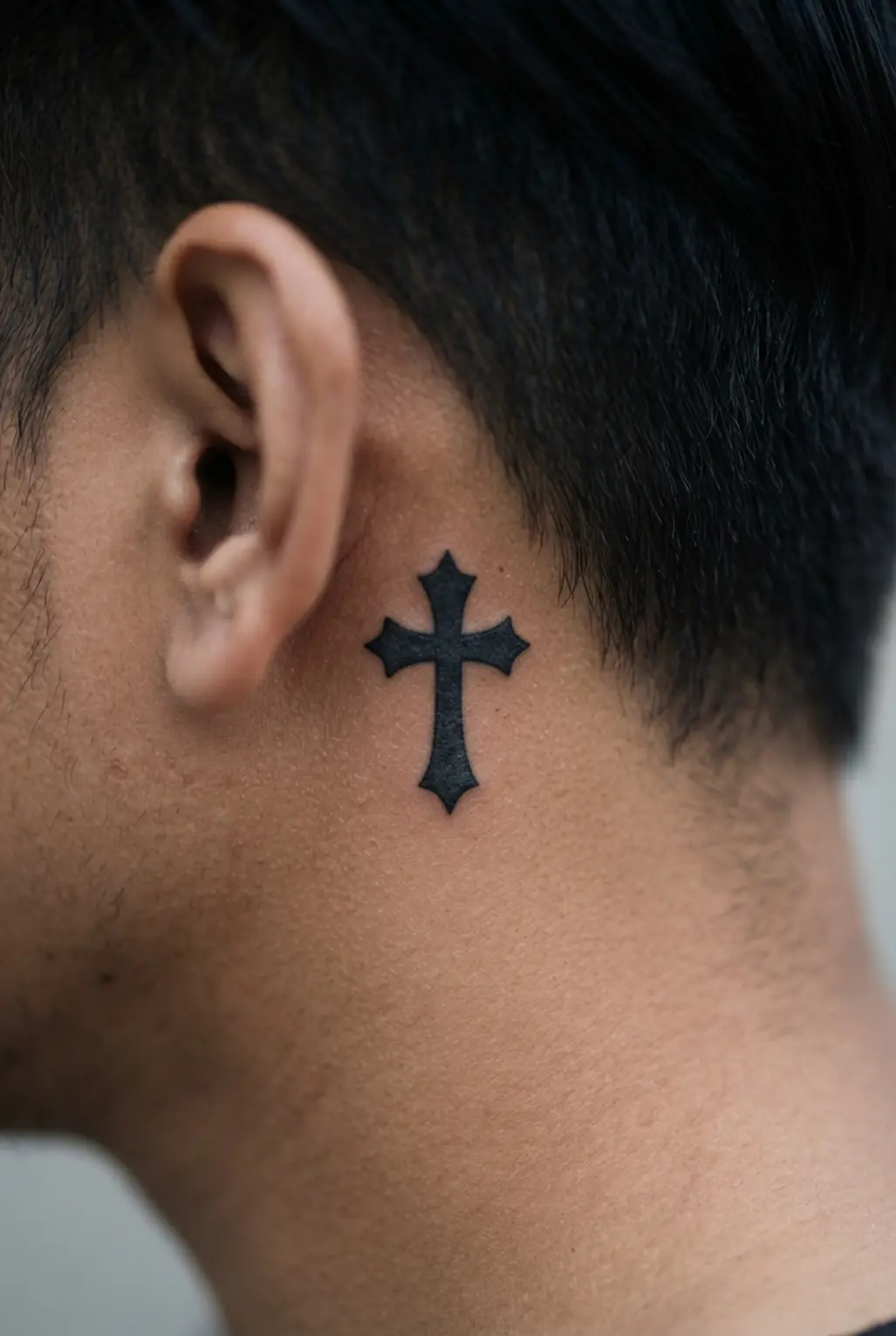 Stylized Gothic cross silhouette tattoo behind the ear, filled with solid black ink and framed by clean pointed edges.