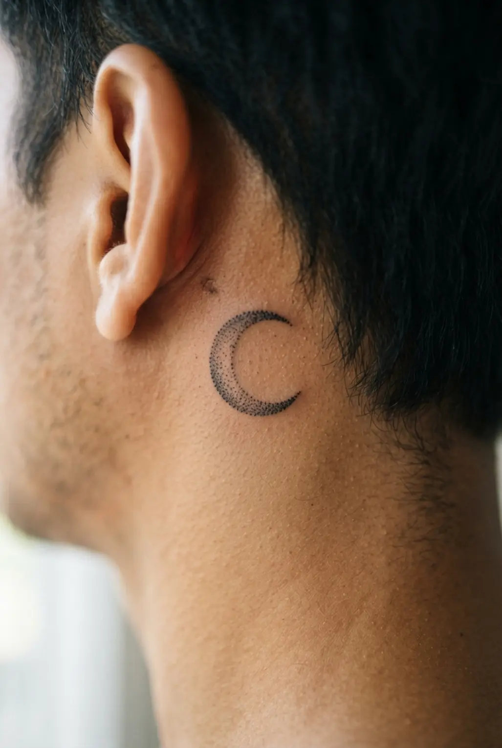 Dotwork crescent moon tattoo behind the ear, using black stippling for a compact masculine design with visible texture.