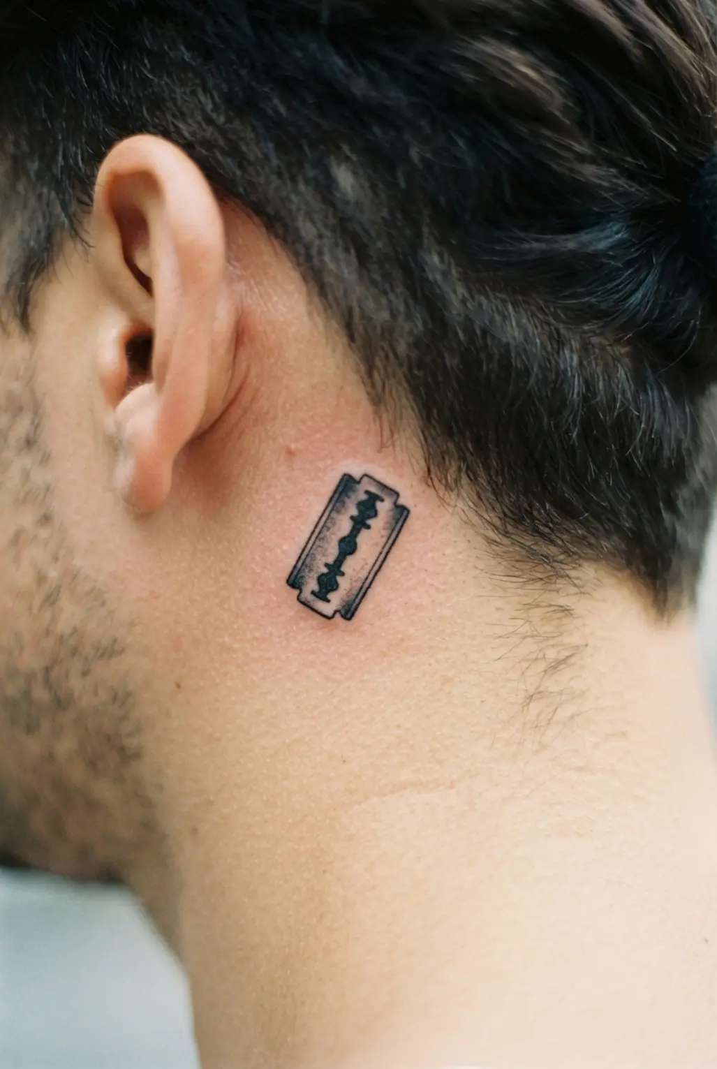 Small razor blade tattoo behind the ear, showing a sharp rectangular shape, dark handle, and clear black outline.