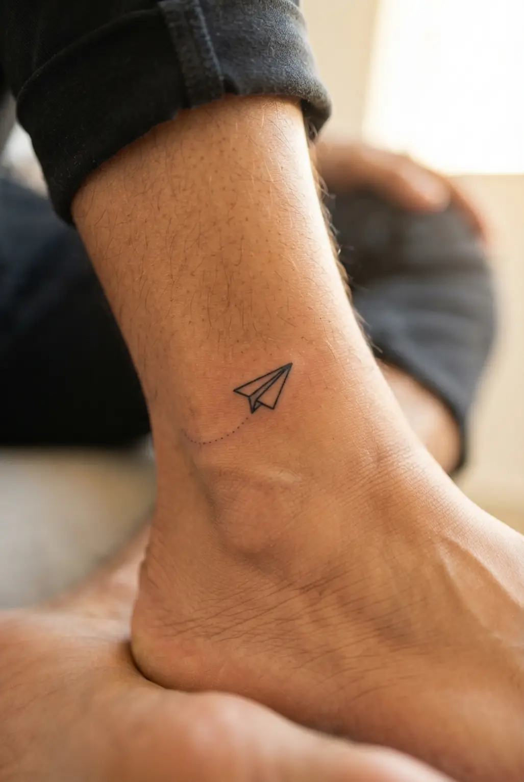 Small paper airplane tattoo near the ankle, drawn in black ink with a simple outline and short flight trail.