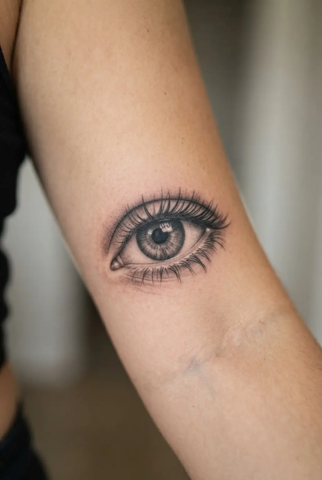 Realistic eye tattoo on the inner bicep with a defined iris, dark lashes, and layered black and gray shading.