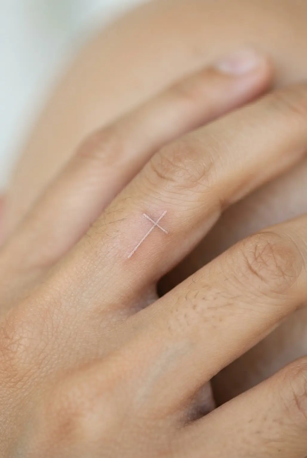 Microscopic white ink cross on a finger, visible as a soft pale symbol with clean lines against natural skin.