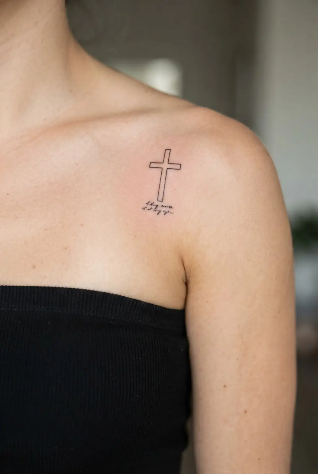 Small scriptural verse cross tattoos on shoulder with fine black lettering beneath a simple outlined cross.