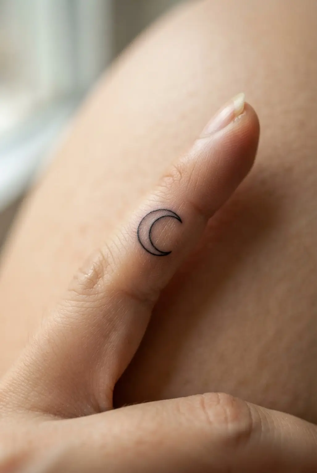 A black fine-line crescent moon rests on a fingertip, showing a clean tiny finger tattoo design.