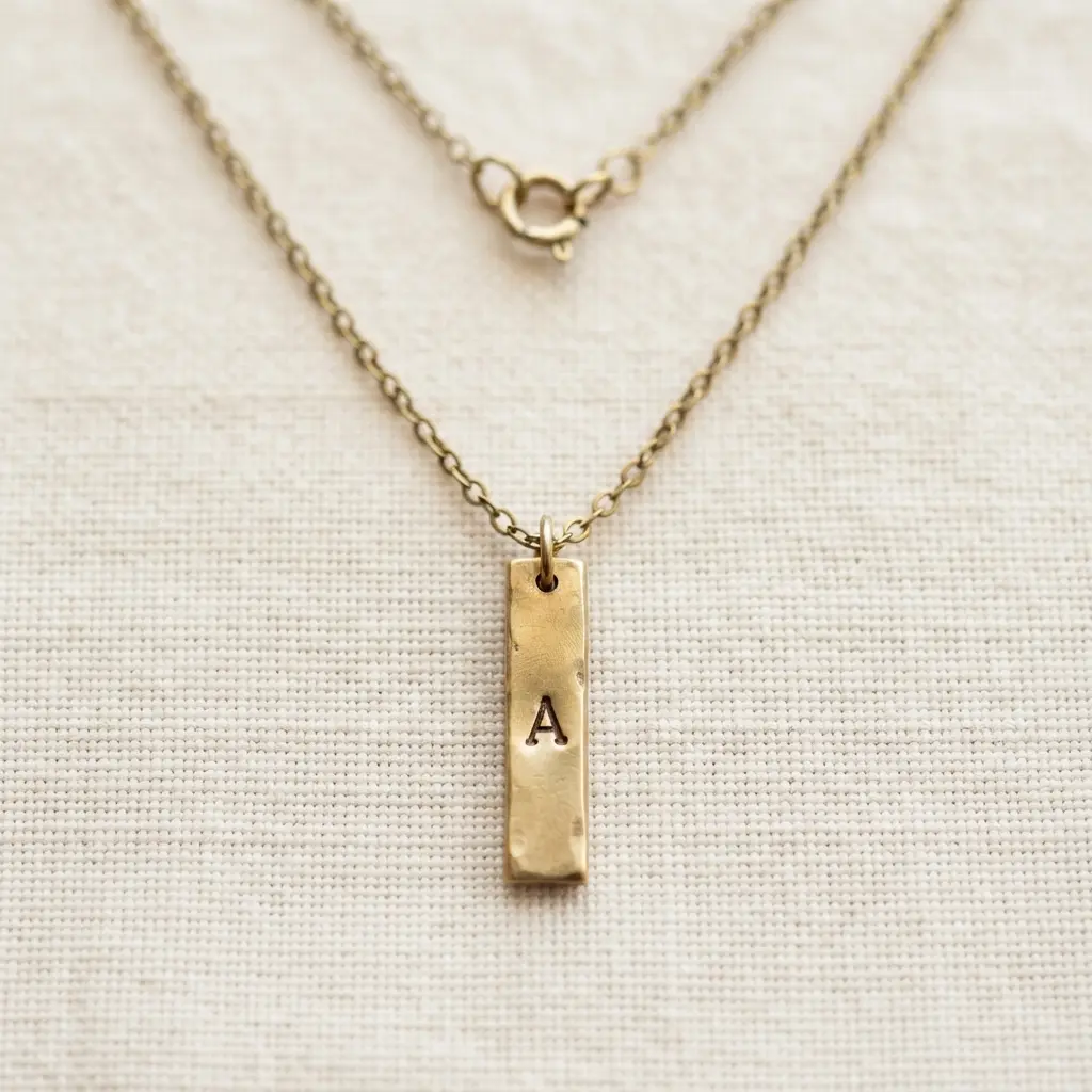 Small stamped metal bar marked with an initial, hanging from a fine chain against a cream background.