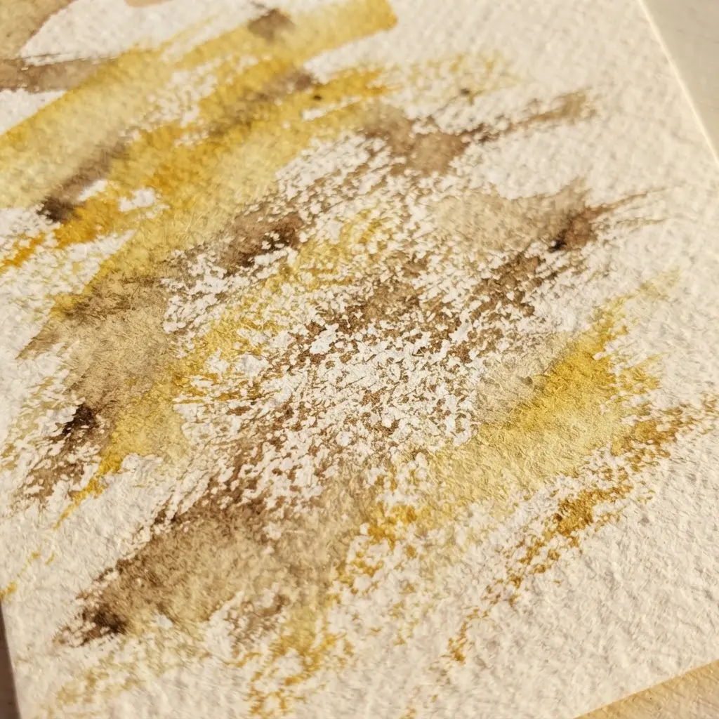 A dry brush leaves broken ochre strokes across textured paper beside a round watercolor brush.