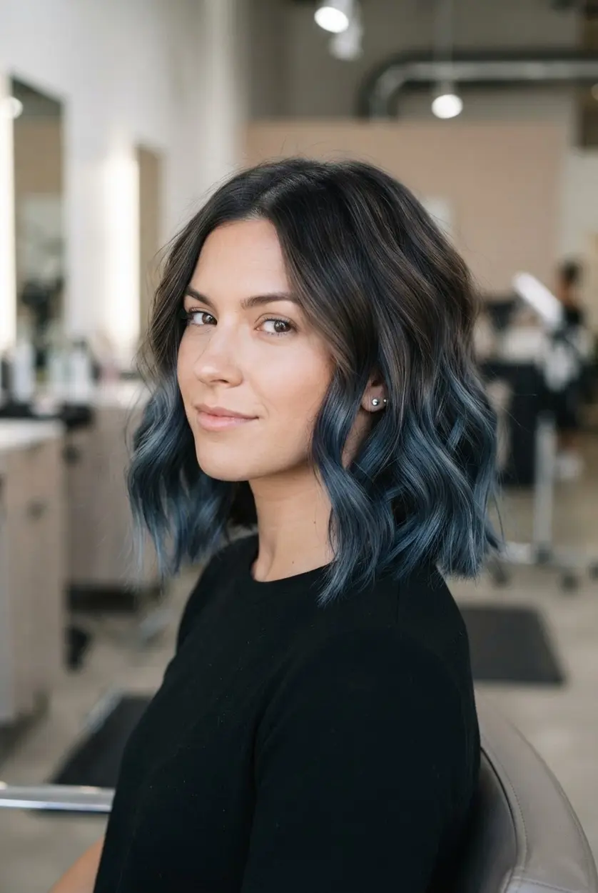 Wavy brunette bob with smoky blue balayage through the lower layers and softly blended dark roots.