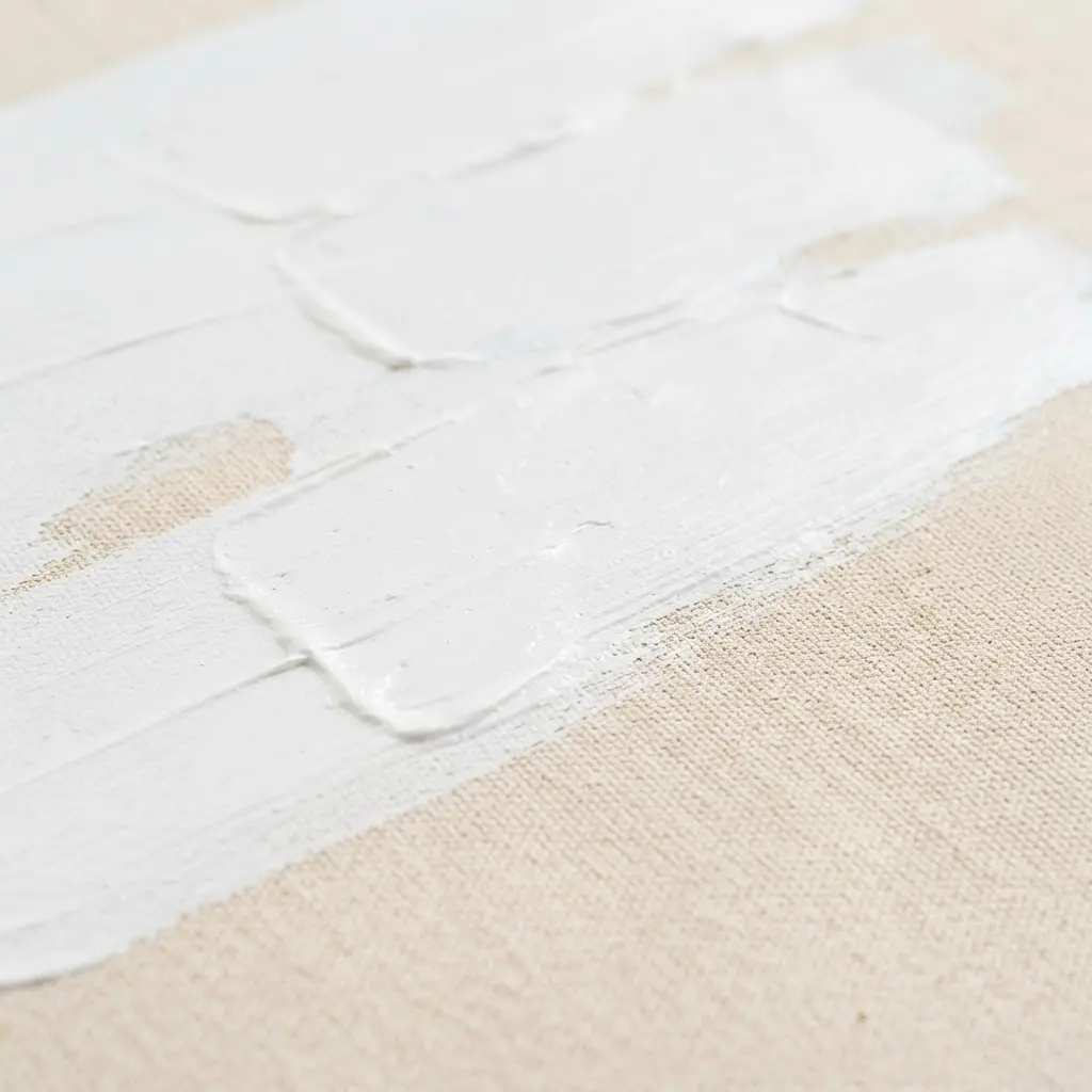 White gesso, a wide brush, and raw cotton canvas sit on a studio table during surface preparation.